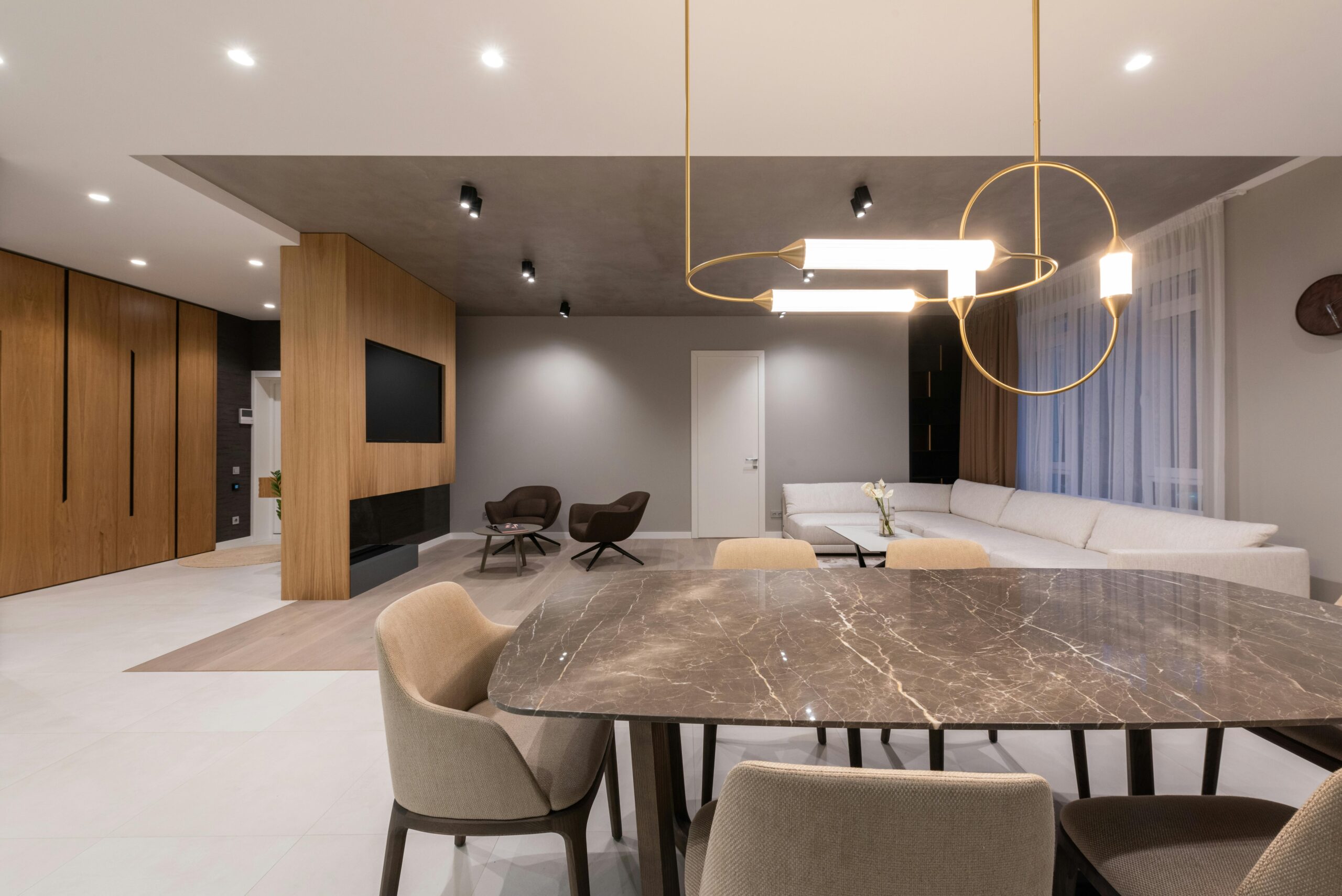 Interior of modern light home with dining table with chairs under lamps hanging on ceiling next to sofa near windows with curtains and armchairs with TV on wall